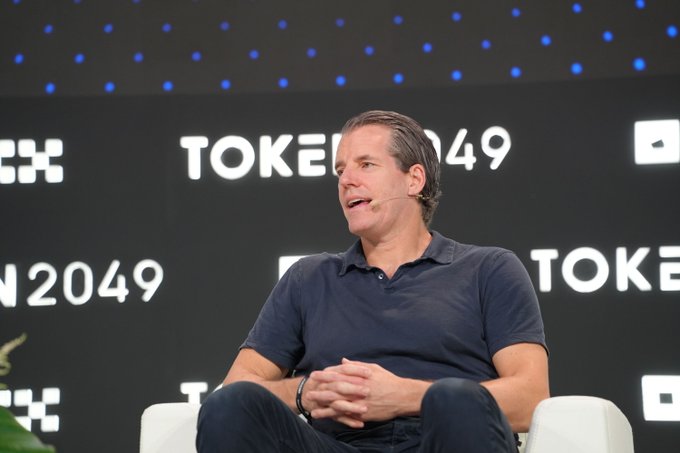 A man in a dark polo shirt speaks at the TOKEN2049 conference, seated in front of a backdrop displaying the event's logo repeatedly.