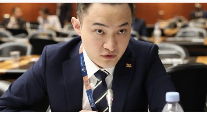 A photo of a man in a dark suit and striped tie wearing a lanyard, seated at what appears to be a formal conference or diplomatic meeting.