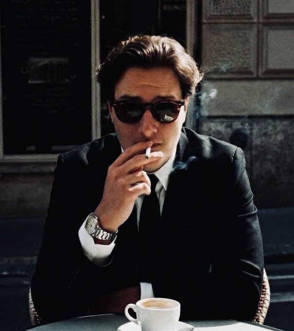 A stylish man wearing dark sunglasses and a black suit with a silver watch smokes a cigarette at an outdoor café table with a coffee cup, reminiscent of a classic film still.