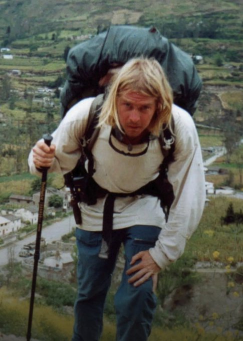 A vintage photograph of a blonde man with a large backpack and trekking pole hiking on a hillside with terraced fields in the background.