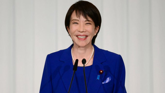 A smiling woman in a blue suit and pearls stands at a podium. Her attire and the Japanese flag pin on her lapel suggest she is a Japanese official, potentially involved in the announcement of state-backed Bitcoin mining.