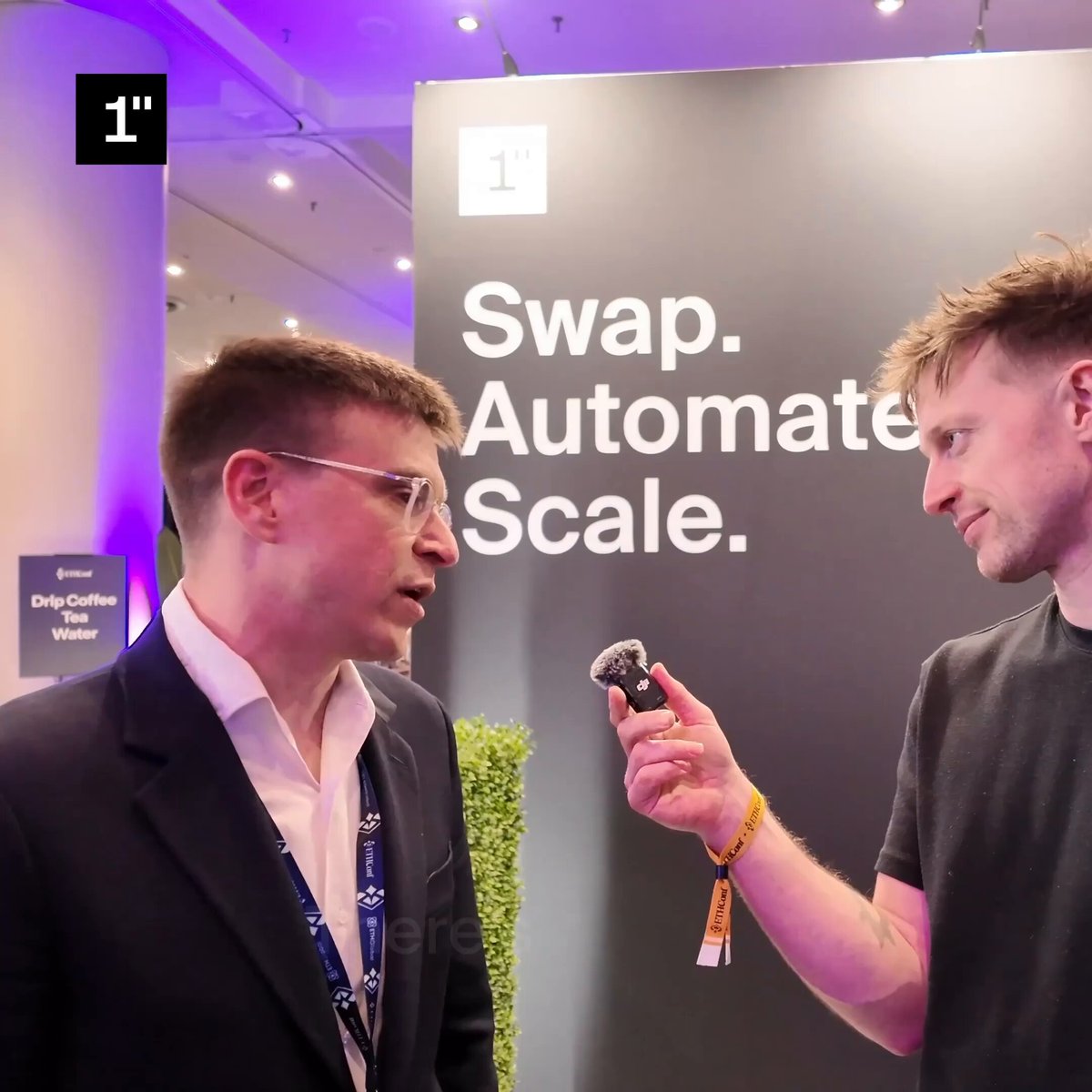The image shows two men standing next to each other, with one man holding a small object while talking to another man who appears to be listening attentively. The background features a sign that reads "Settlement creates stablecoin balances. Then they become balance sheets that have to be managed." This suggests that the conversation might be related to blockchain technology or financial management in general.