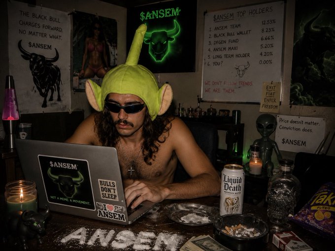 A shirtless man wearing a green Shrek-style hat with ears and sunglasses works at a laptop covered in $ANSEM stickers, surrounded by $ANSEM cryptocurrency memorabilia, posters, and meme references including 'Bulls Don't Sleep' and 'Liquid Death'.