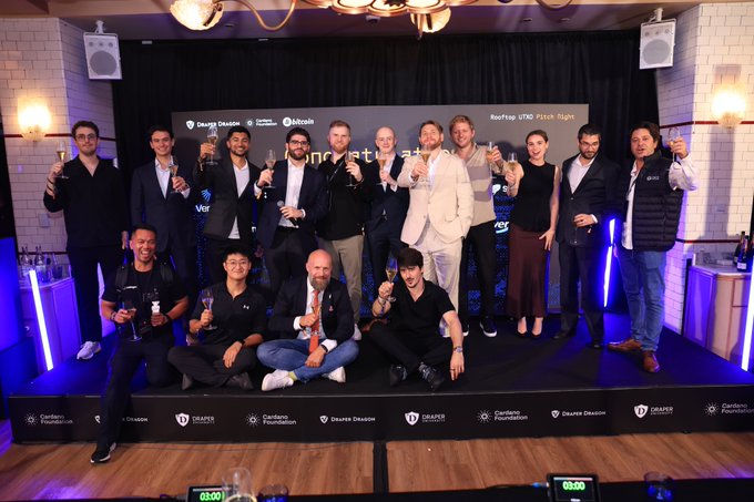 A group photo of approximately 15 people posing on a stage at an event, holding drinks and giving thumbs up. The backdrop displays logos including 'Design Dashio', 'Cardano Foundation', and 'bitcoin', suggesting a Cardano hackathon or crypto-related event.