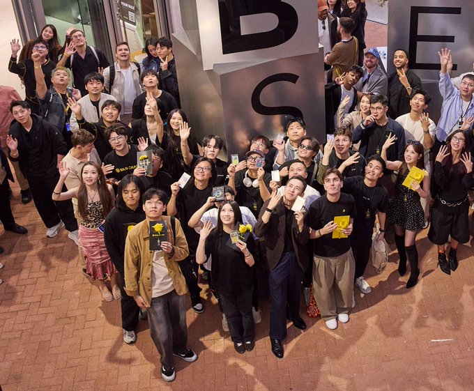 A large group of people pose together for a photo in front of a wall with large letters spelling 'DESIGN', holding various items including yellow books.