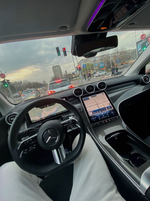 A driver sits in a Mercedes-Benz (identifiable by the steering wheel logo) with the large infotainment screen displaying a navigation map, stopped at a red light on a city street. The luxurious car interior paired with the motivational "let's make $100k this August" message symbolizes an aspirational mindset toward financial success at the start of the new month.