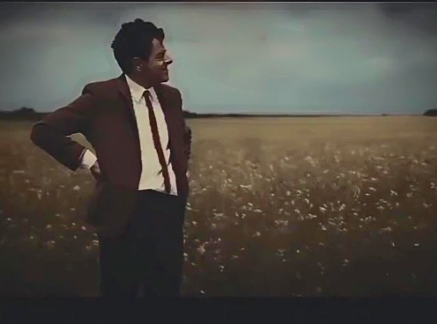 A cinematic, slightly grainy shot of a man standing in a vast, grassy field. He is wearing a dark reddish-brown suit with a tie, looking off to the side against an overcast sky, reminiscent of the film 'The Conformist'.
