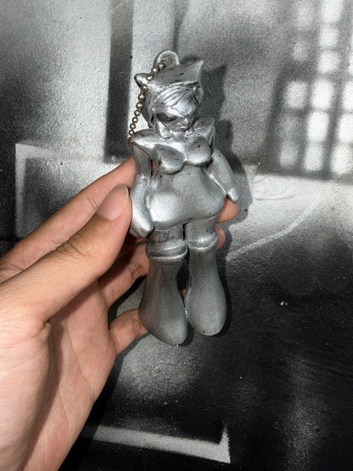 urn of a silver metal figurine with a white face and black hair, wearing a pink dress and red shoes, held by a hand against a black background.