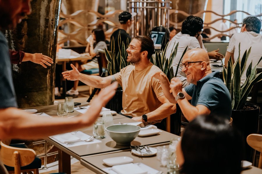 Two men enthusiastically greet each other with outstretched arms at a bustling restaurant table, smiling broadly in a lively dining atmosphere.