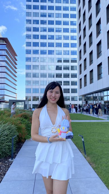 A smiling young woman in a white dress holds a small plush toy while posing outdoors with modern office buildings in the background.