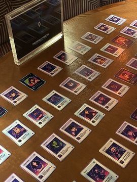 A spread of Monad Cards collectible trading cards is laid out on a wooden table next to a digital display, with the speaker jokingly asking if this arrangement is a sign to host another "Monad Cards" private dinner.