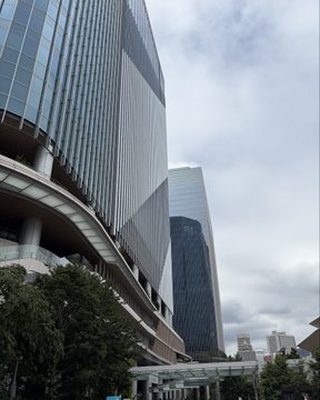 A modern glass skyscraper rises against a cloudy sky, with curved walkways and greenery at its base, likely near the LAWSON Takana Gateway City store where the JPYC payment proof-of-concept experiment is taking place.