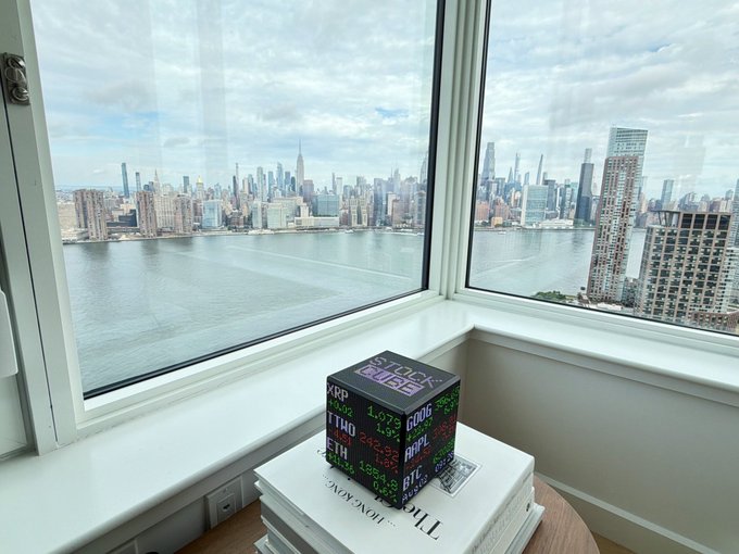 A cube-shaped LED display showing stock tickers like SPX, XOM, and ETH sits on a stack of books by a window, overlooking a sweeping view of the Manhattan skyline and East River.