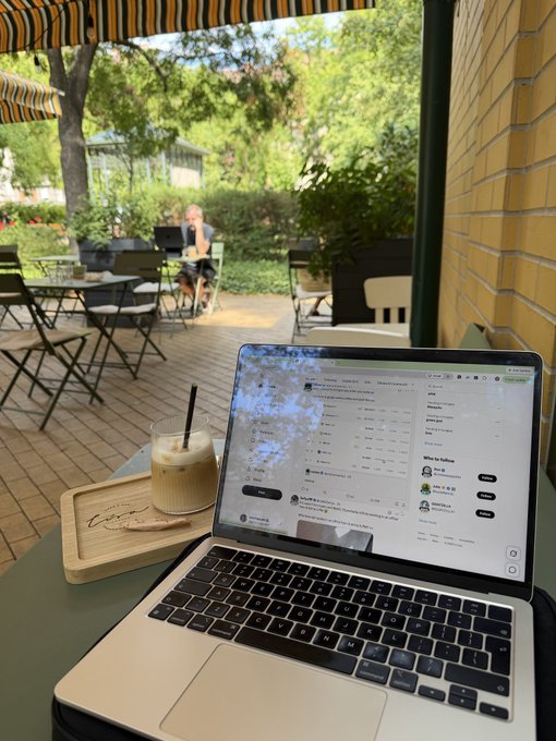 A laptop sits on an outdoor café table displaying what appears to be a social media feed, alongside an iced coffee drink on a wooden tray, with a person seated at another table in the background.