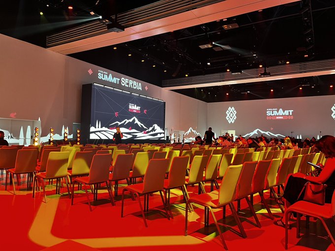 Photo of the Solana Summit Serbia conference venue, showing rows of empty chairs facing a stage with the summit branding on screens and a mountainous backdrop.