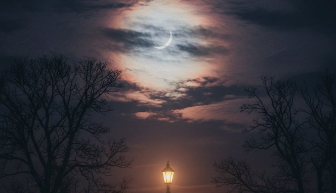 The image features a street lamp with a crescent moon behind it, creating a beautiful nighttime scene. The sky above has clouds, adding depth to the overall atmosphere of the photo. There are no people or other objects visible in the image, making it an interesting and captivating view for those who appreciate the beauty of nature during the night hours.