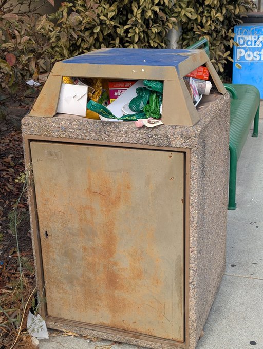 An overflowing public trash bin on a sidewalk, with garbage and debris spilling out of its top, next to a green bench and hedge.