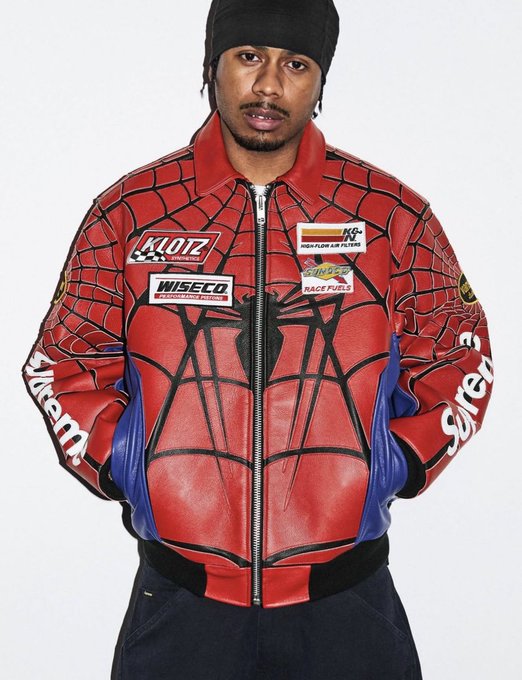 A man wearing a black beanie models a red and blue leather jacket featuring a Spider-Man-inspired web design and racing sponsor patches, including "KLOTZ," "WISECO," "Sunoco Race Fuels," and "KG Hightower Air Filters." The sleeves display "Supreme" branding in white lettering.