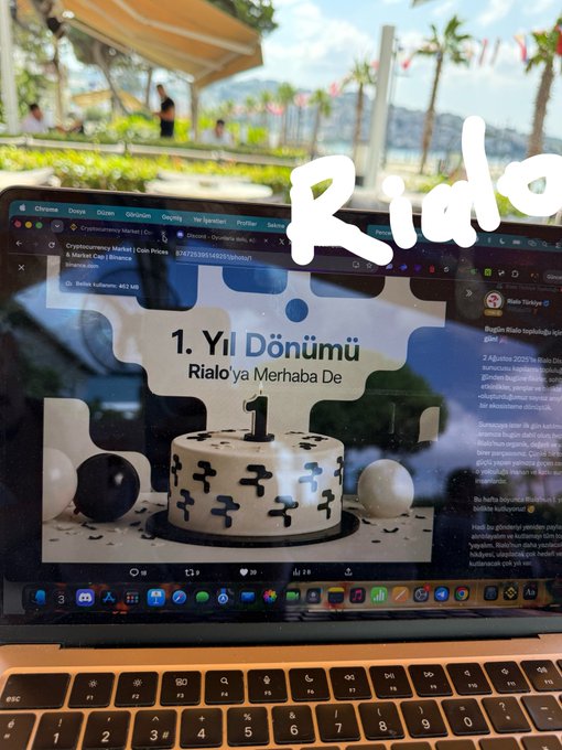 A laptop screen displays a '1. Yıl Dönümü / Rialo'ya Merhaba De' 1st anniversary webpage with a cake graphic, viewed outdoors with the word 'Rialo' added as overlay text.