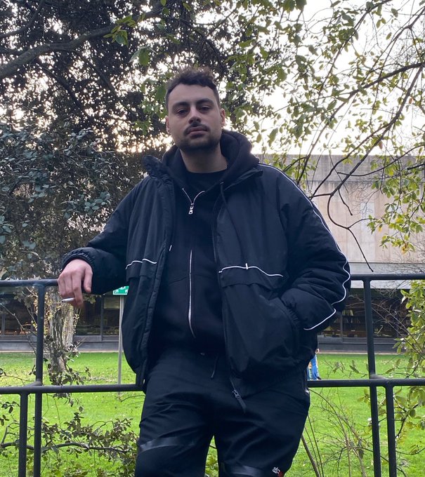 A young man wearing a black tracksuit and puffer jacket stands outdoors by a fence with trees in the background.