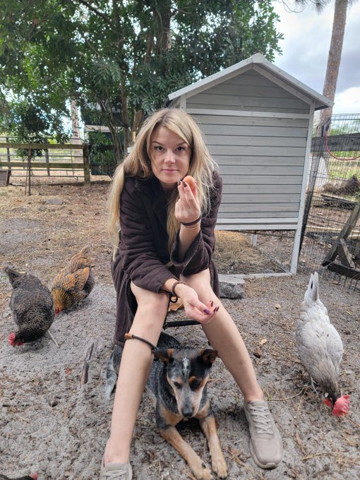 A blonde woman in a brown robe sits on a stool in a yard, holding her hand up toward the camera, surrounded by chickens and with a small dog resting at her feet.
