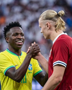 Photo of two soccer players shaking hands on the field, one wearing a yellow Brazil jersey and the other in a red jersey with blonde hair in a bun.