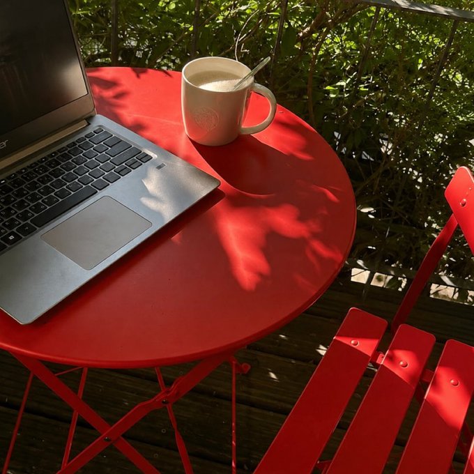 A silver Acer laptop sits open on a round red metal patio table next to a white Starbucks mug with a spoon, accompanied by a matching red folding chair on a wooden deck with greenery in the background.