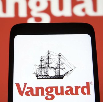 The image shows a smartphone displaying the Vanguard logo, which features a black illustration of a tall ship above the company name in red text. The phone is set against a red background with partial "anguard" lettering visible, emphasizing the company's brand identity in connection with the announcement that Vanguard is hiring a Head of Bitcoin and Digital Assets.
