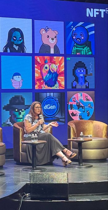 A woman speaking into a microphone on stage at an 'NFT' conference, seated in front of a large screen displaying various NFT collections including Bored Apes, Pudgy Penguins, and a dGen logo.