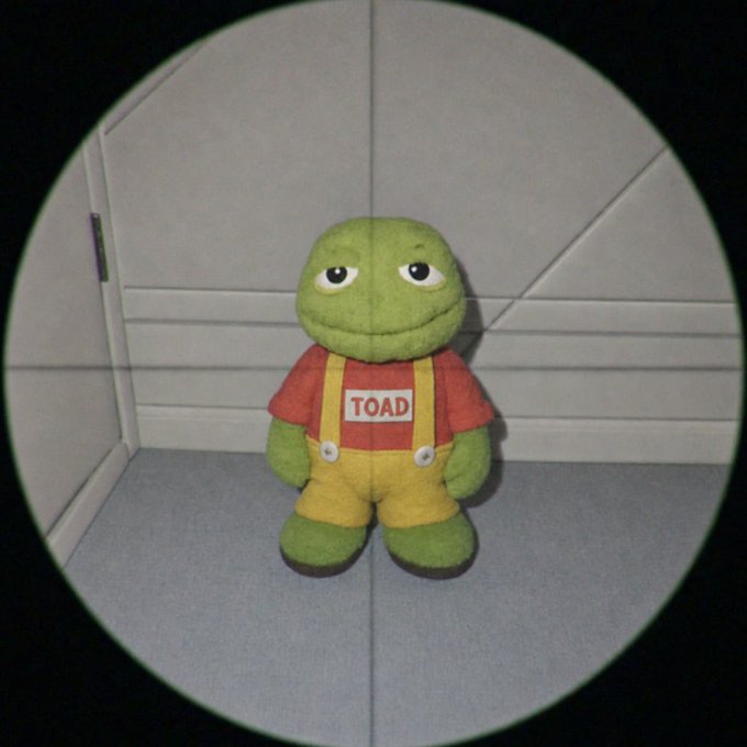 A plush toy resembling Pepe the Frog wearing a red shirt with a "TOAD" name tag and yellow overalls, viewed through a circular sniper scope or camera lens with crosshairs.