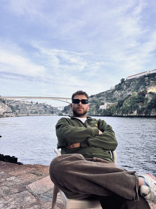 A man in sunglasses and a green jacket sits by the water with a bridge in the background, looking relaxed. The image is related to the post asking for $SOL addresses, implying the man might be the one distributing them.