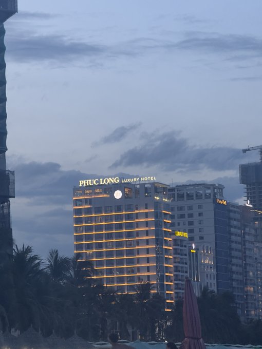 An evening view of the Phuc Long Luxury Hotel building with its illuminated sign glowing against a dusky sky, alongside other high-rise developments. The hotel's prominent branding is featured in the scene, suggesting a focus on hospitality sector developments or tourism-related market activity in Vietnam.