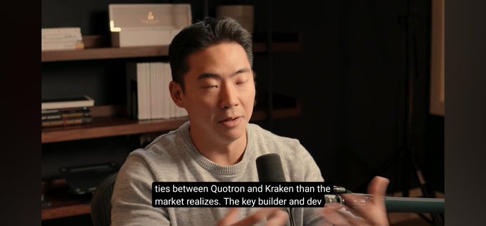 A man in a gray sweater speaks into a microphone during what appears to be a podcast or interview, with subtitles reading 'ties between Quotron and Kraken than the market realizes. The key builder and dev'.