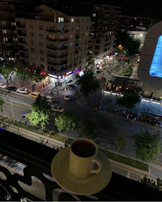 The image shows a cup of coffee sitting on top of a balcony railing, overlooking a city street below at night. The scene captures the essence of a peaceful evening with the warm glow of the city lights illuminating the surroundings. The photo appears to be taken from an elevated position, providing a unique perspective that combines urban life and personal tranquility.