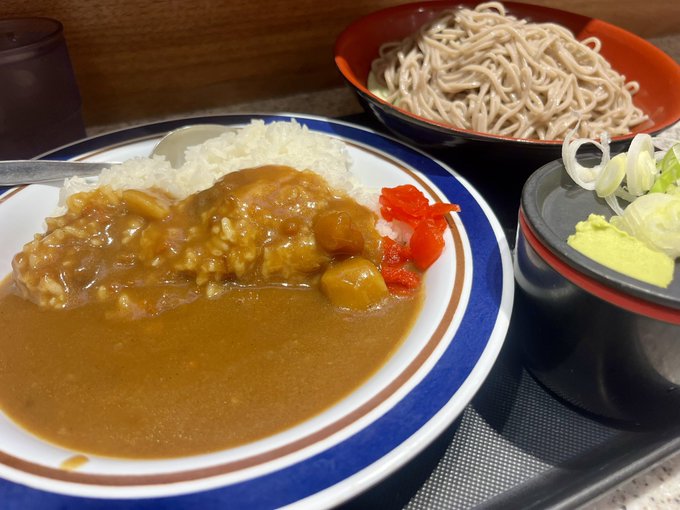 A plate of Japanese curry rice sits alongside a bowl of cold soba noodles and a small side of condiments, reflecting the post-farewell meal mentioned in the caption after finishing a job interview. The accompanying hashtags reference picking up a new smartphone at Apple Marunouchi and checking STEPN, a move-to-earn app.