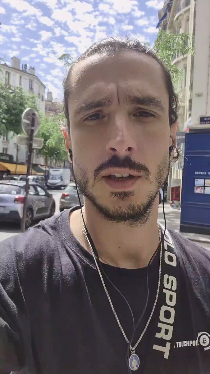 The image features a man wearing earbuds while standing outside of a building with a car parked nearby. The man has a serious expression, possibly contemplating something or reacting to an unexpected event. There are also several cars visible in the background, indicating that he is likely in an urban setting. The post about the Freedomia card and Bitcoin addresses seems unrelated to the image itself but could be related to the man's current situation if there was any connection between his earbuds and the card.