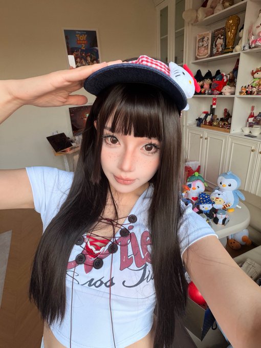 A selfie of a young woman with long dark hair and bangs wearing a light blue corset-style top with red lace-up details and a black hat with a red checkered bow and a Hello Kitty charm, standing in a room filled with plush toys and collectibles.