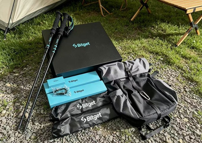 A Bitget-branded mountaineering gift box containing trekking poles, a black duffel bag, and blue accessory boxes sits on gravel beside a tent, complementing the post about receiving the crypto exchange's 8th anniversary gift set symbolizing rising success.
