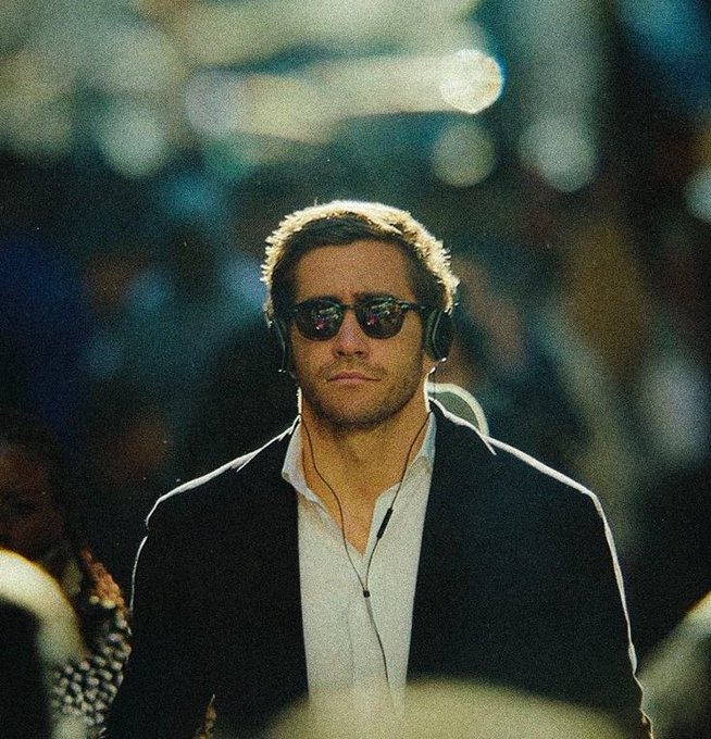 A man wearing dark sunglasses, headphones, and a dark blazer over a white shirt, standing in a dimly lit environment with blurred lights in the background.