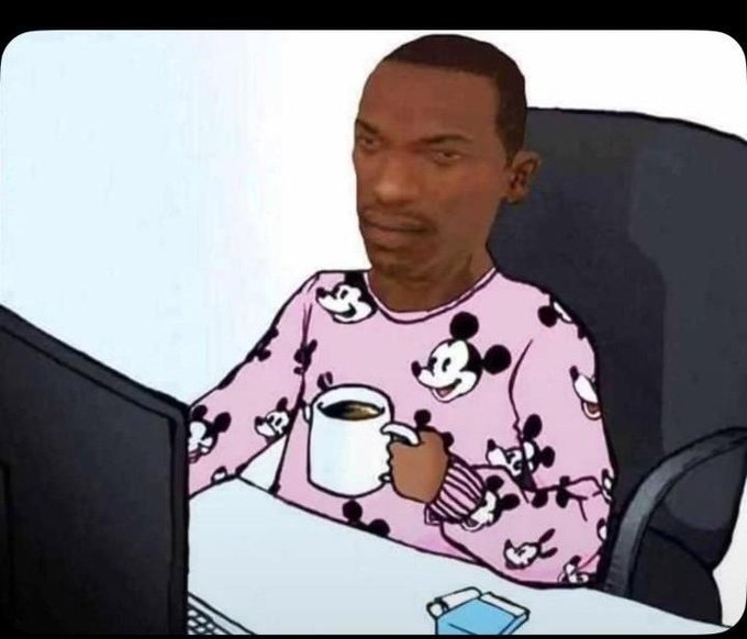 A man wearing pink Mickey Mouse pajamas sits at a desk holding a coffee mug and looking at a computer monitor, with a pack of cigarettes nearby.