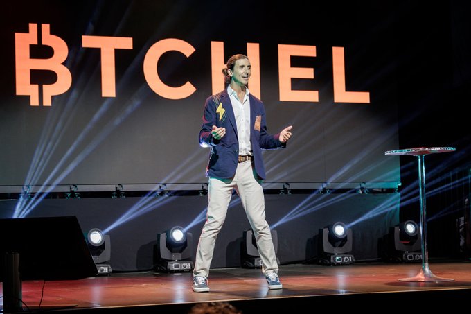 A man with a ponytail, wearing a blue jacket and white pants, speaking on a stage in front of a screen displaying the word "BITCHEL" alongside a Bitcoin logo.
