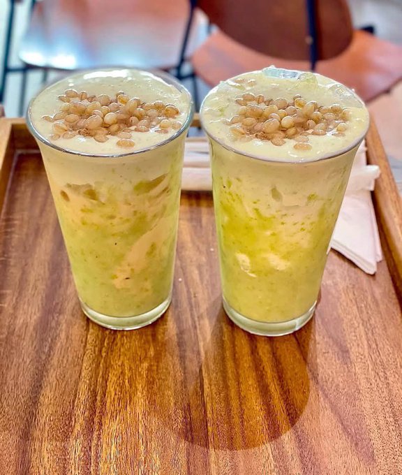 Two tall glasses of a green blended beverage topped with crunchy grains or puffed cereal, served on a wooden tray with napkins.