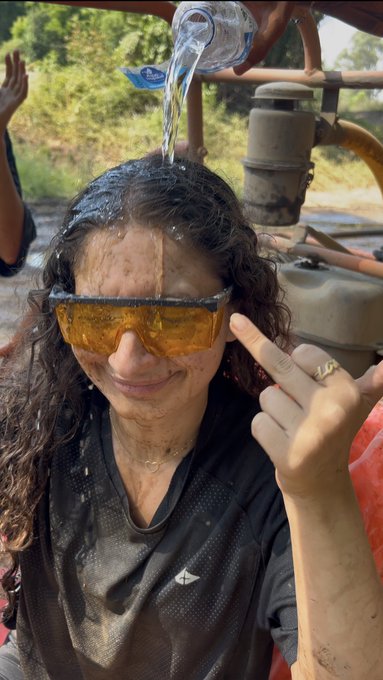 A woman wears orange ski goggles while water is poured over her head, capturing the messy aftermath of what appears to be a "I got my sticker" tradition—her wet hair and grinning peace sign suggest she's just earned her novelty decal, covered in mud and laughter.