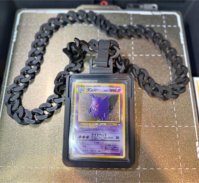 A black chain necklace displayed in a presentation case, featuring a rectangular pendant that holds a Japanese Pokémon trading card depicting the purple ghost-type Pokémon Gengar.