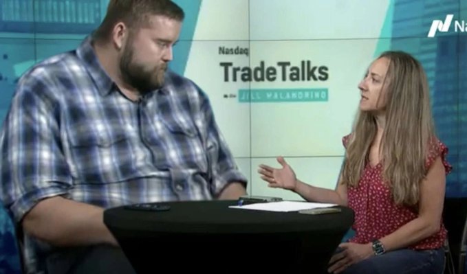 A man in a blue plaid shirt and a woman in a red polka-dot top sit at a table during a 'Nasdaq TradeTalks with Jill Malandrino' interview segment.