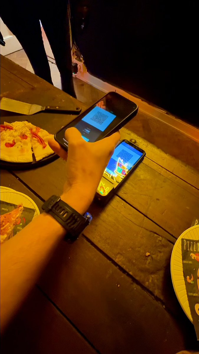 The image shows a person holding up their phone displaying a pizza order for Bitcoin Pizza day. The person's hand is visible, while the cell phone screen captures the attention of those around them. In the background, there are two pizzas on plates placed on a dining table.