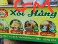 A store sign that reads "XỐI HÀNG" with red text on a yellow background, featuring images of food items and additional Vietnamese text below.