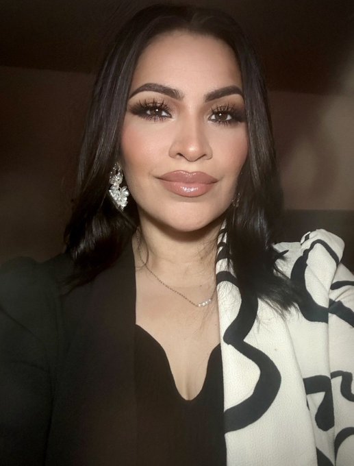A woman with long dark hair, dramatic makeup, and statement floral earrings poses for a selfie wearing a black top and a black-and-white patterned scarf, appearing polished and ready for the day as the caption wishes followers a good morning and asks about their crypto investments.