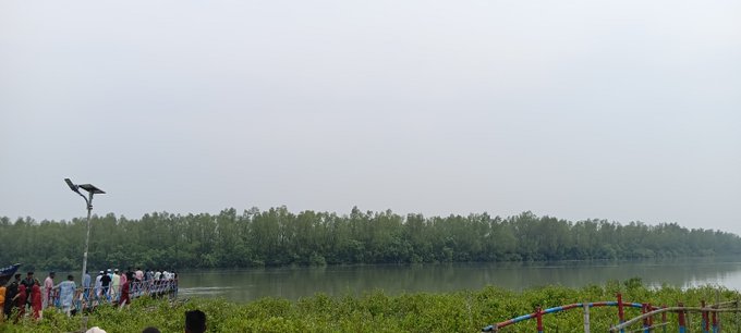 A misty, overcast waterside scene showing a group of people gathered at the edge of a river or waterway, with a bird (possibly an eagle) perched on a pole, mangrove trees lining the opposite shore, and a colorful railing in the foreground.