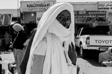 A black and white photograph of a young man wearing a traditional white head wrap and robe, standing outdoors with vehicles and Arabic signage visible in the background.